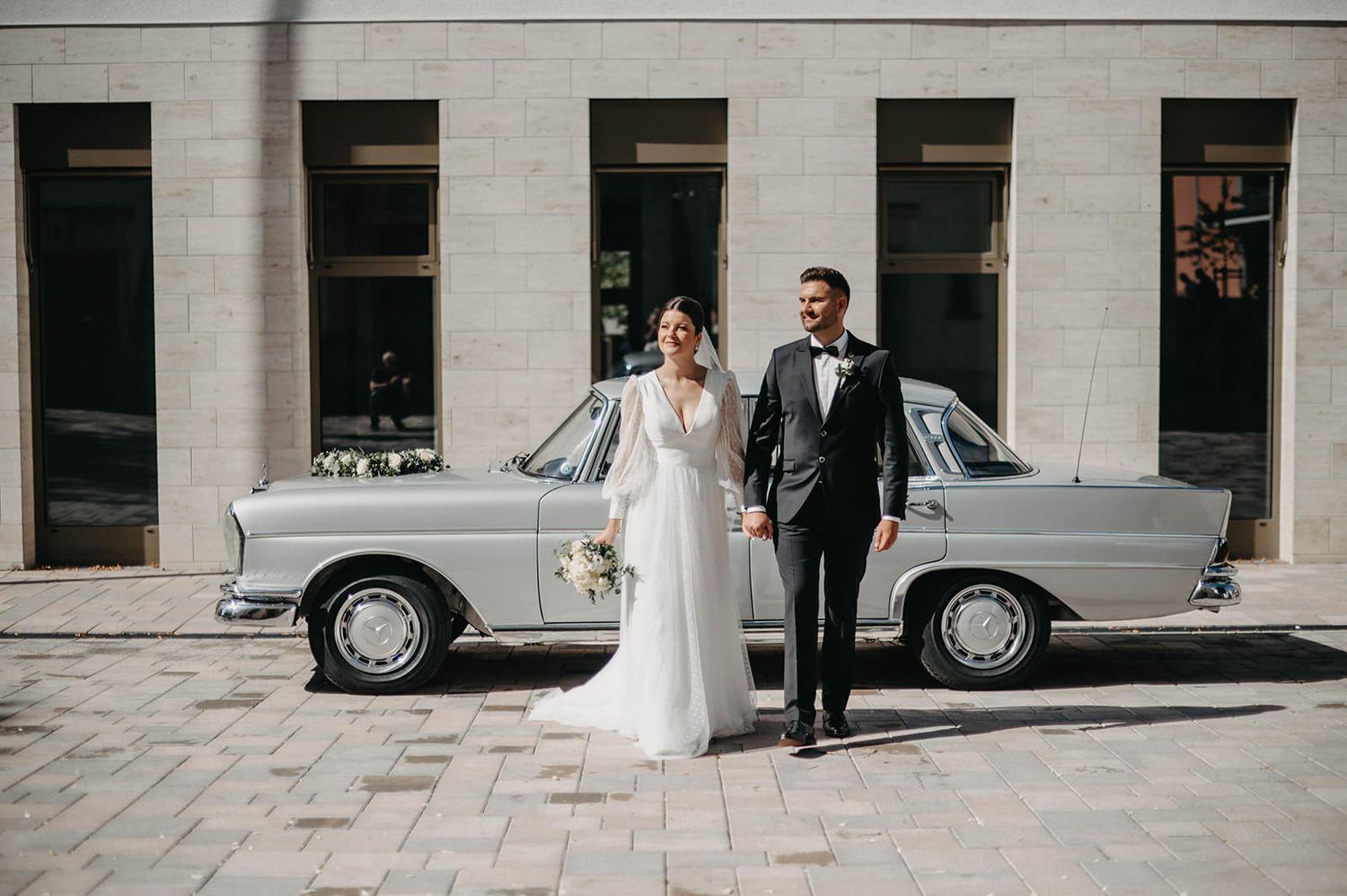 A newlywed couple stands beside a vintage silver Mercedes-Benz on a cobblestone street in front of a stone building.