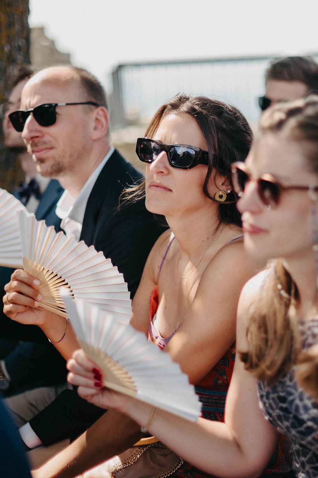 Wedding guests with sunglasses hold paper fans while sitting outdoors during a sunny ceremony.