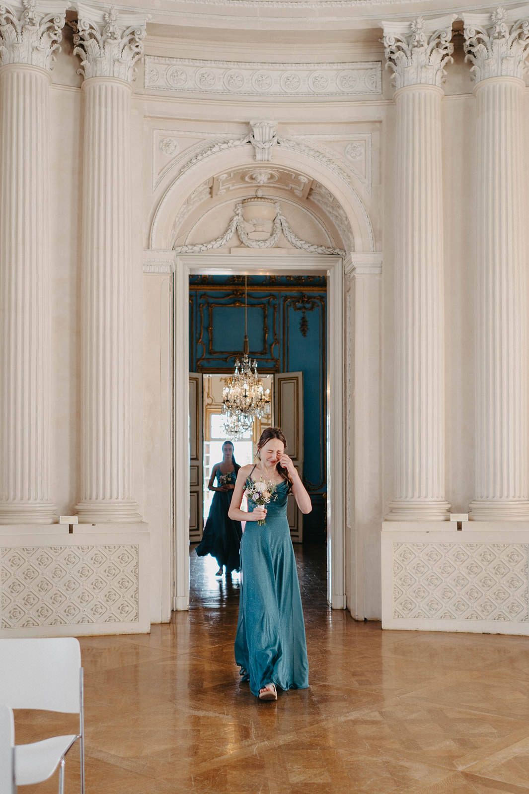 Elegant ballroom interior with ornate white columns and archway featuring a teal door and polished hardwood floors.