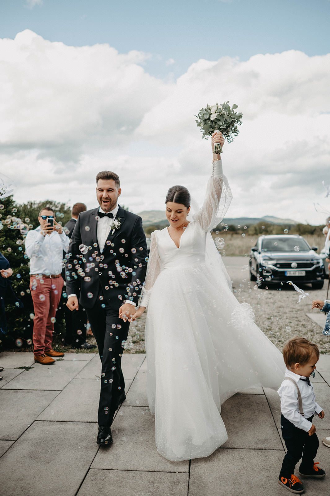 Wedding guests shower confetti on newlyweds during outdoor celebration with cloudy sky backdrop.
