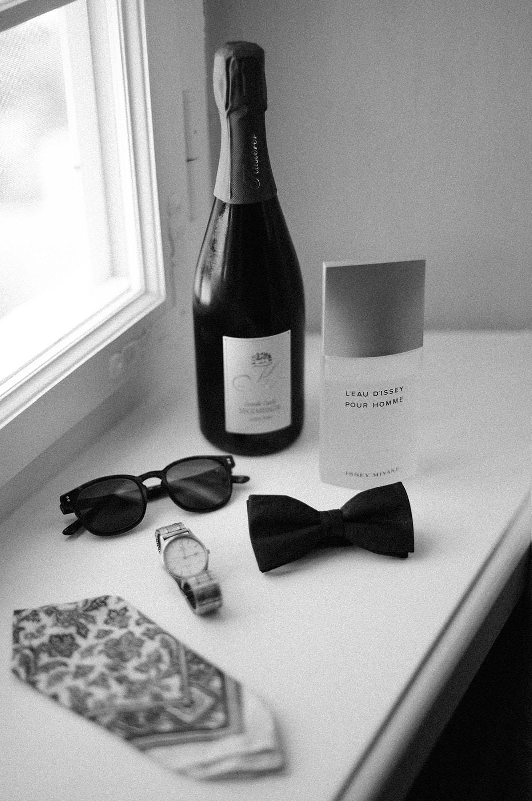 Wedding accessories including champagne bottle, bow tie and sunglasses arranged on white windowsill.