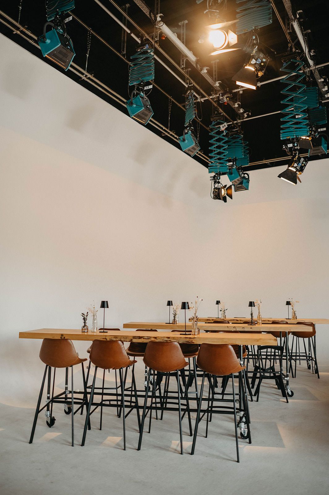 Modern studio space with wooden tables and black metal chairs arranged in rows under industrial lighting and turquoise fixtures.
