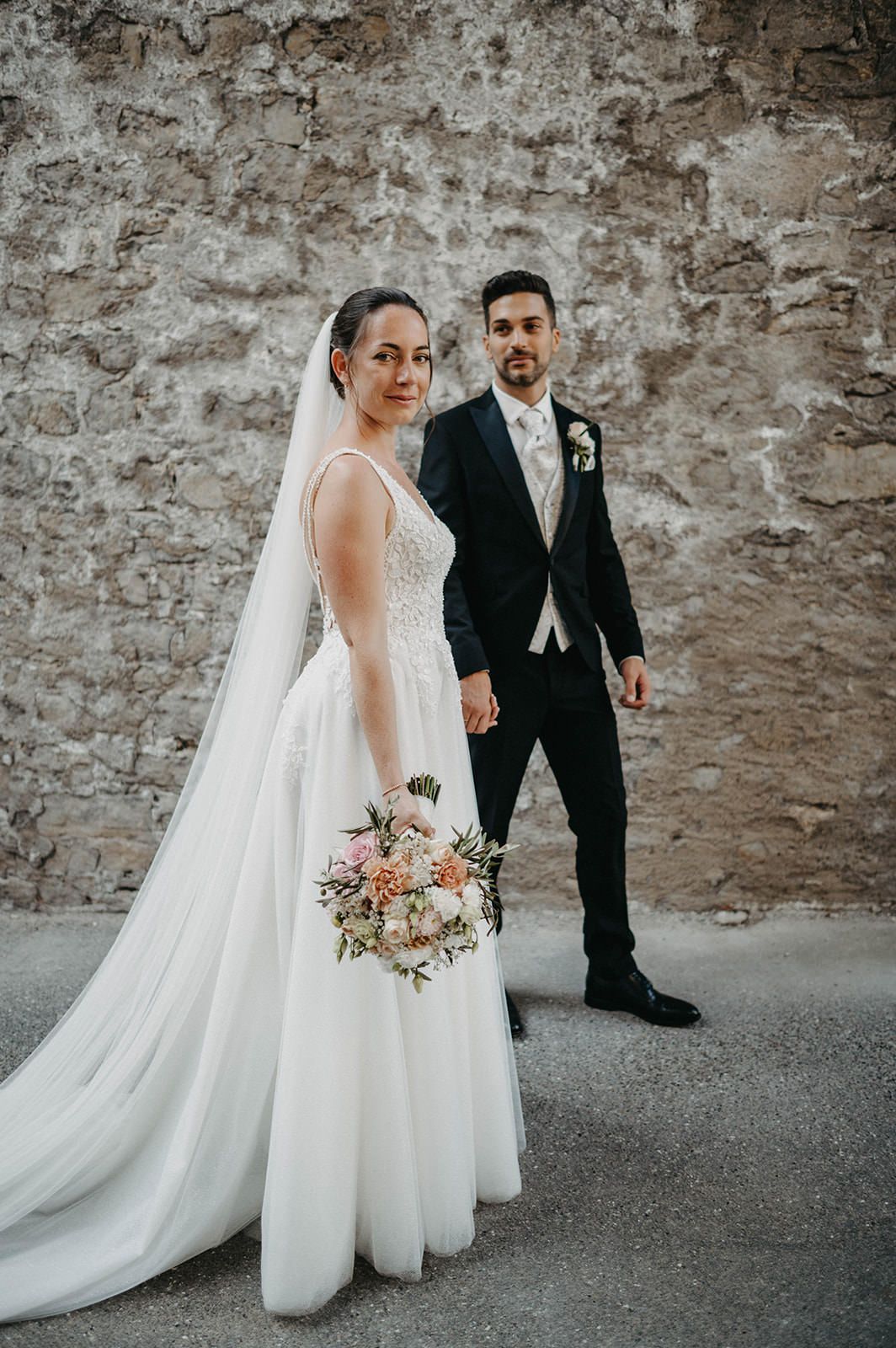 A bride and groom walk together against a rustic stone wall backdrop during their wedding ceremony.