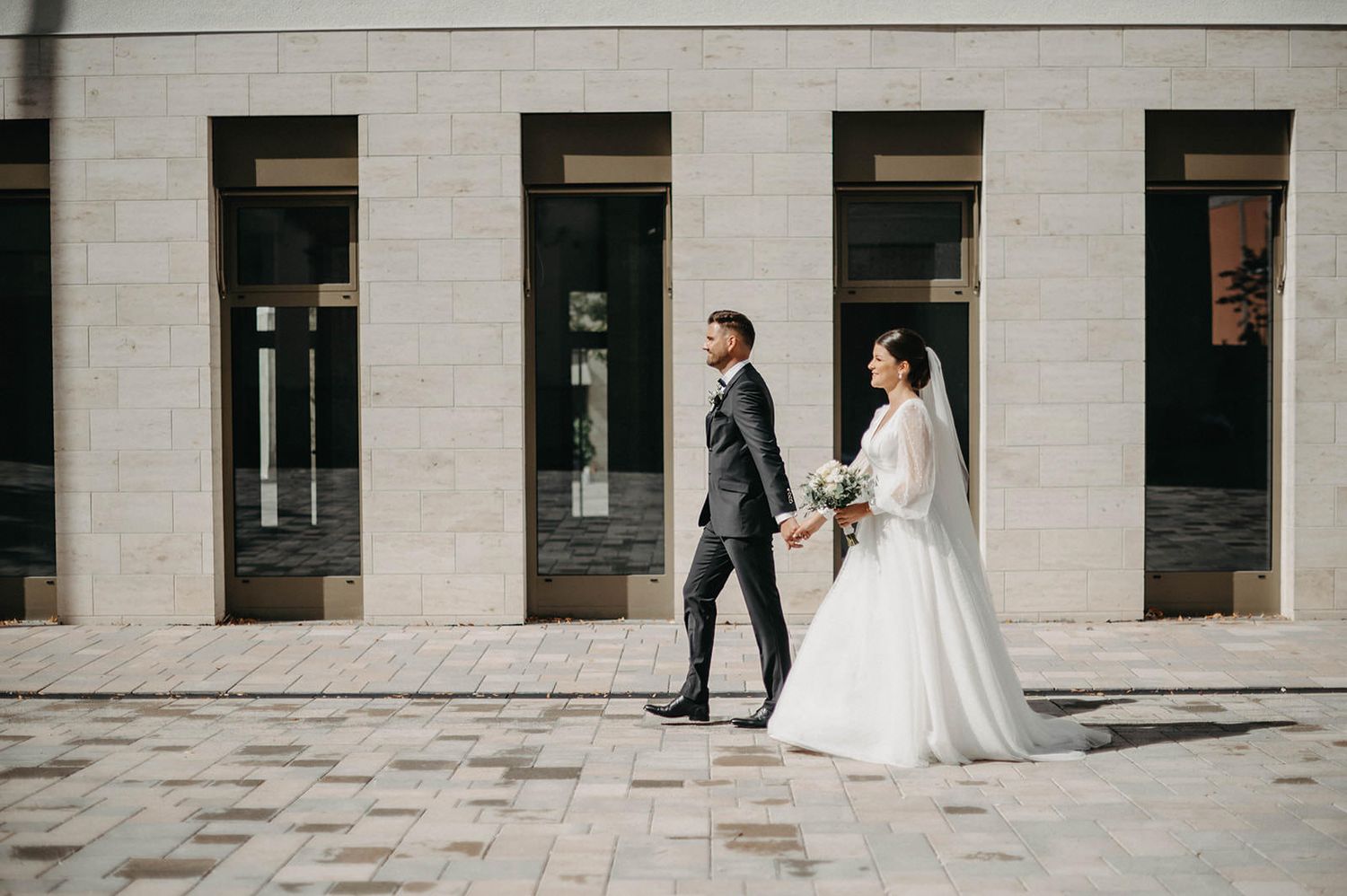 Bride and groom walk together through an elegant stone colonnade with repeating architectural columns.
