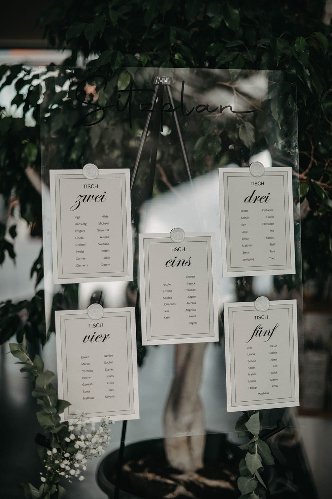 White wedding seating chart cards with German numbers hanging on display against a dark background with greenery.