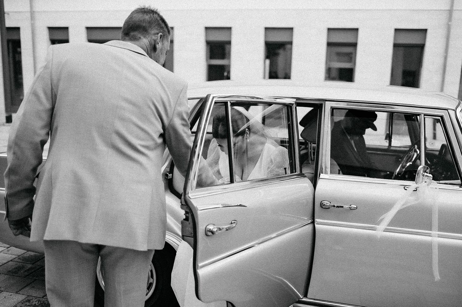 A person in a light colored suit opens the door of a vintage car in a black and white photograph.