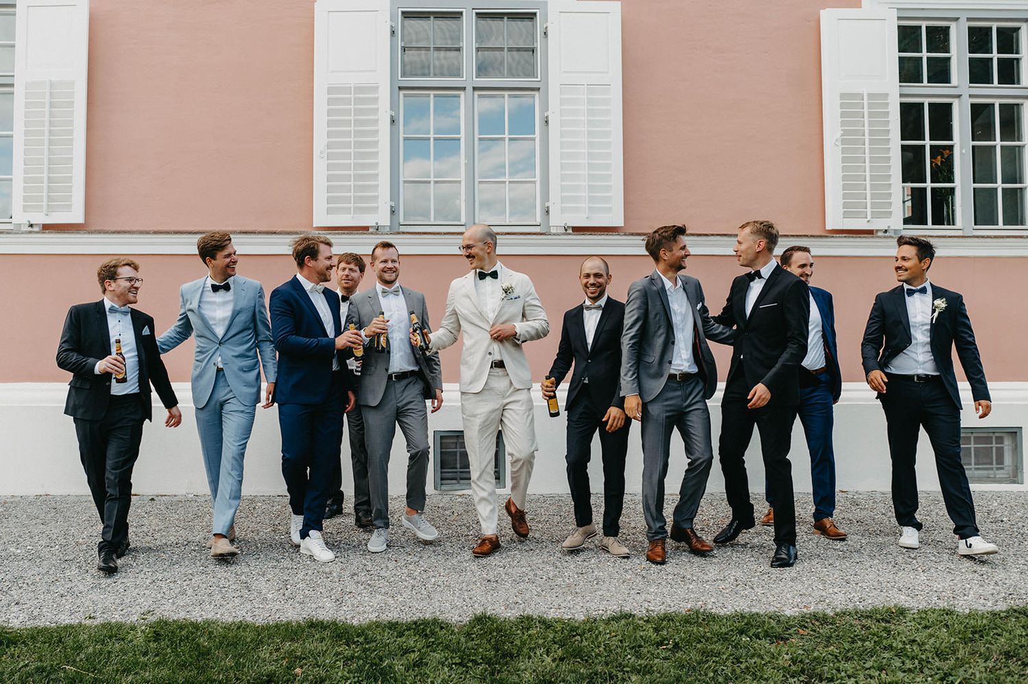 A group of people in formal attire lean against a white building wall in a candid wedding party photo.