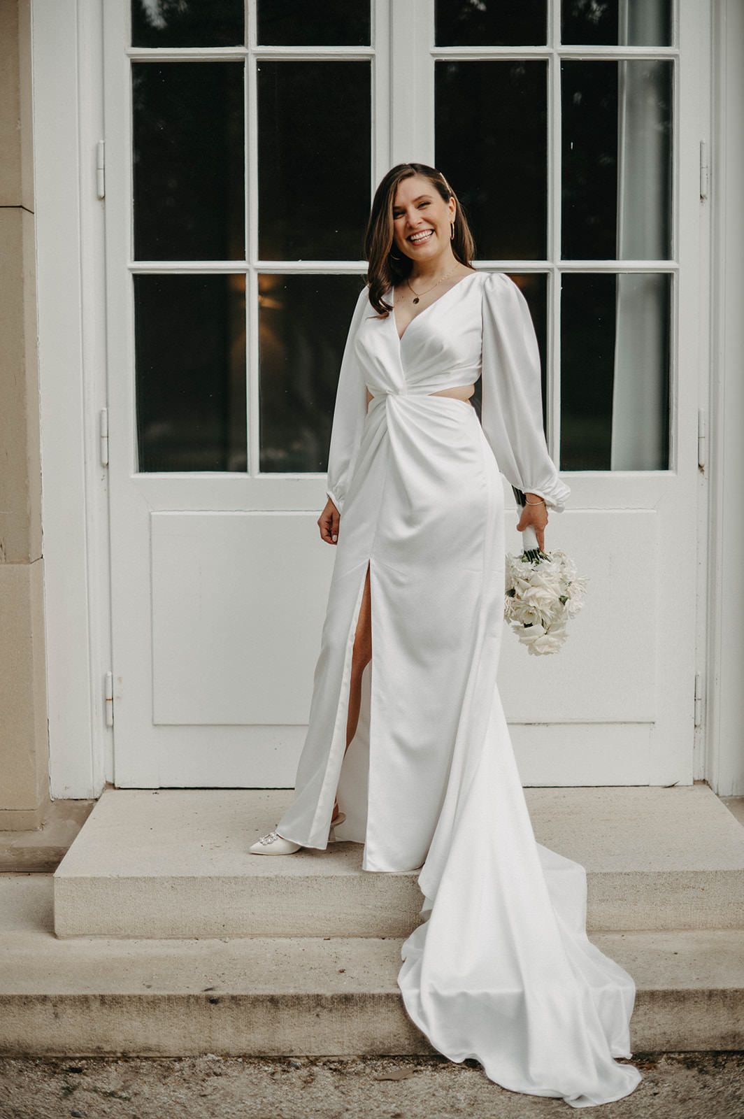 Elegant bride in long white dress with balloon sleeves and side slit poses on stone steps by French doors.
