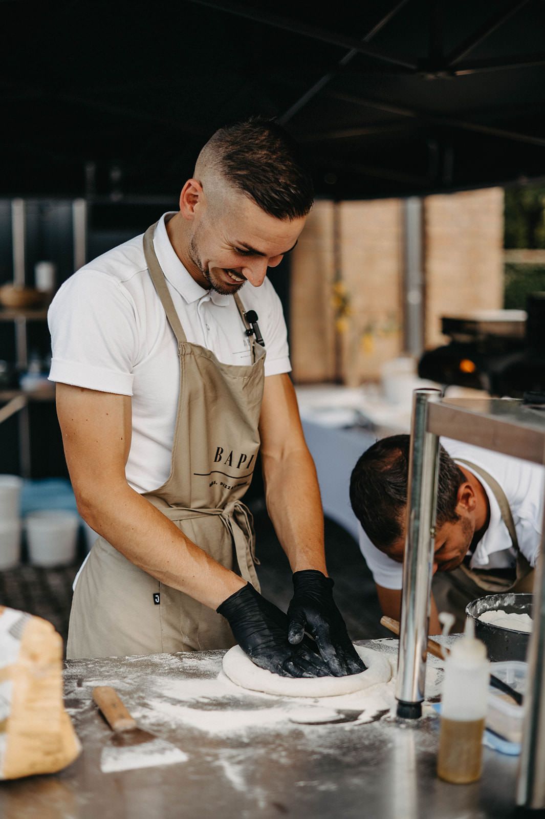 Chef in white shirt and tan apron carefully shapes dough on a floured surface in professional kitchen.
