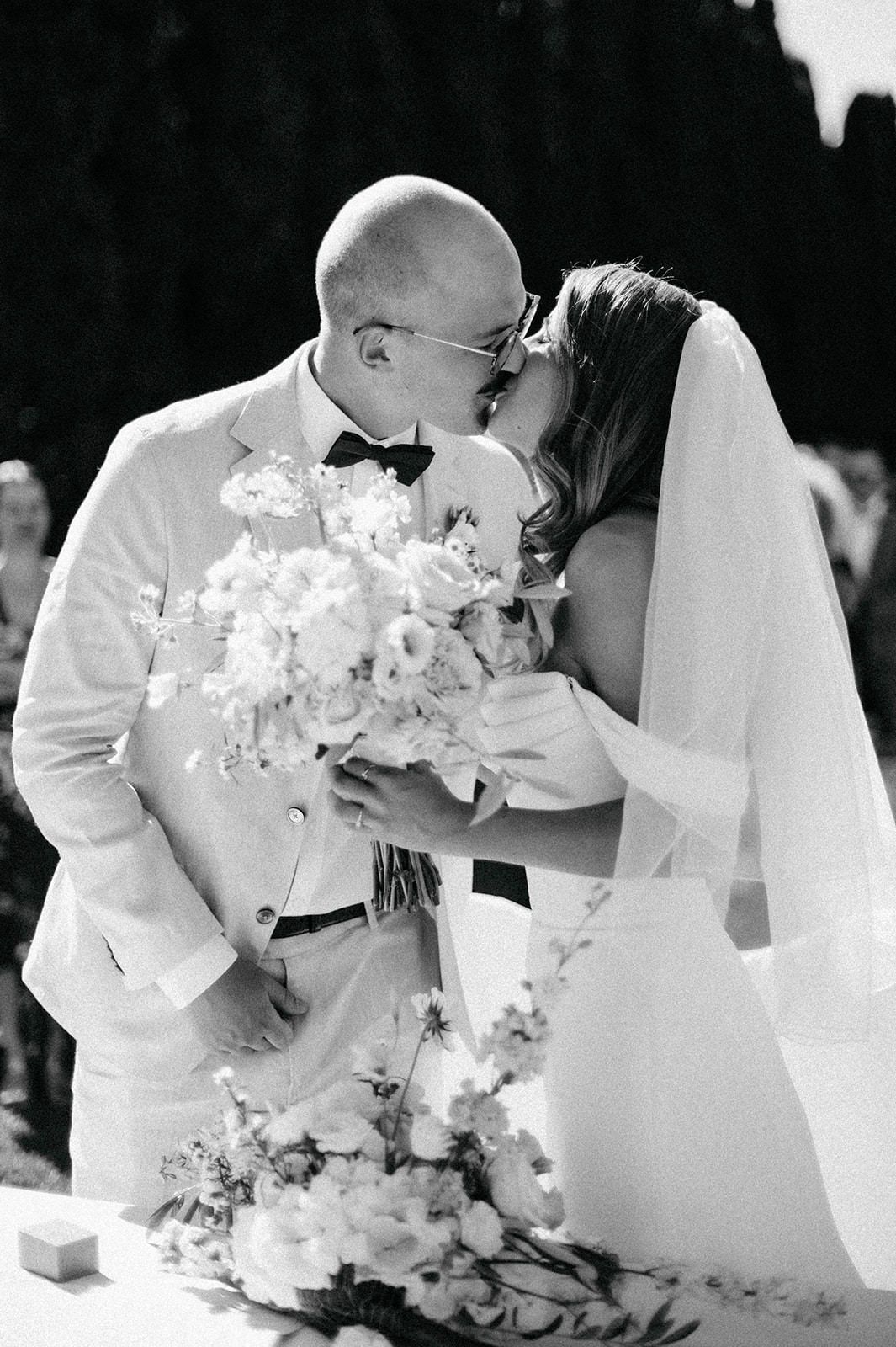 Black and white wedding photo series showing couple sharing intimate kiss behind elegant white orchid bouquet.
