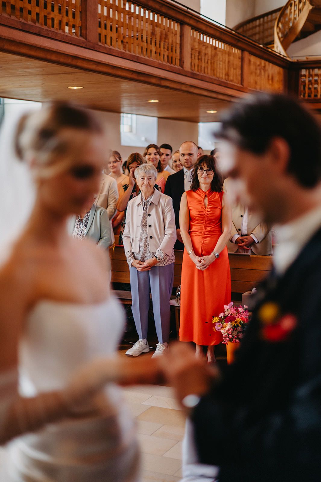 Wedding guests stand in church pews during an emotional ceremony with warm lighting and ornate wooden details.