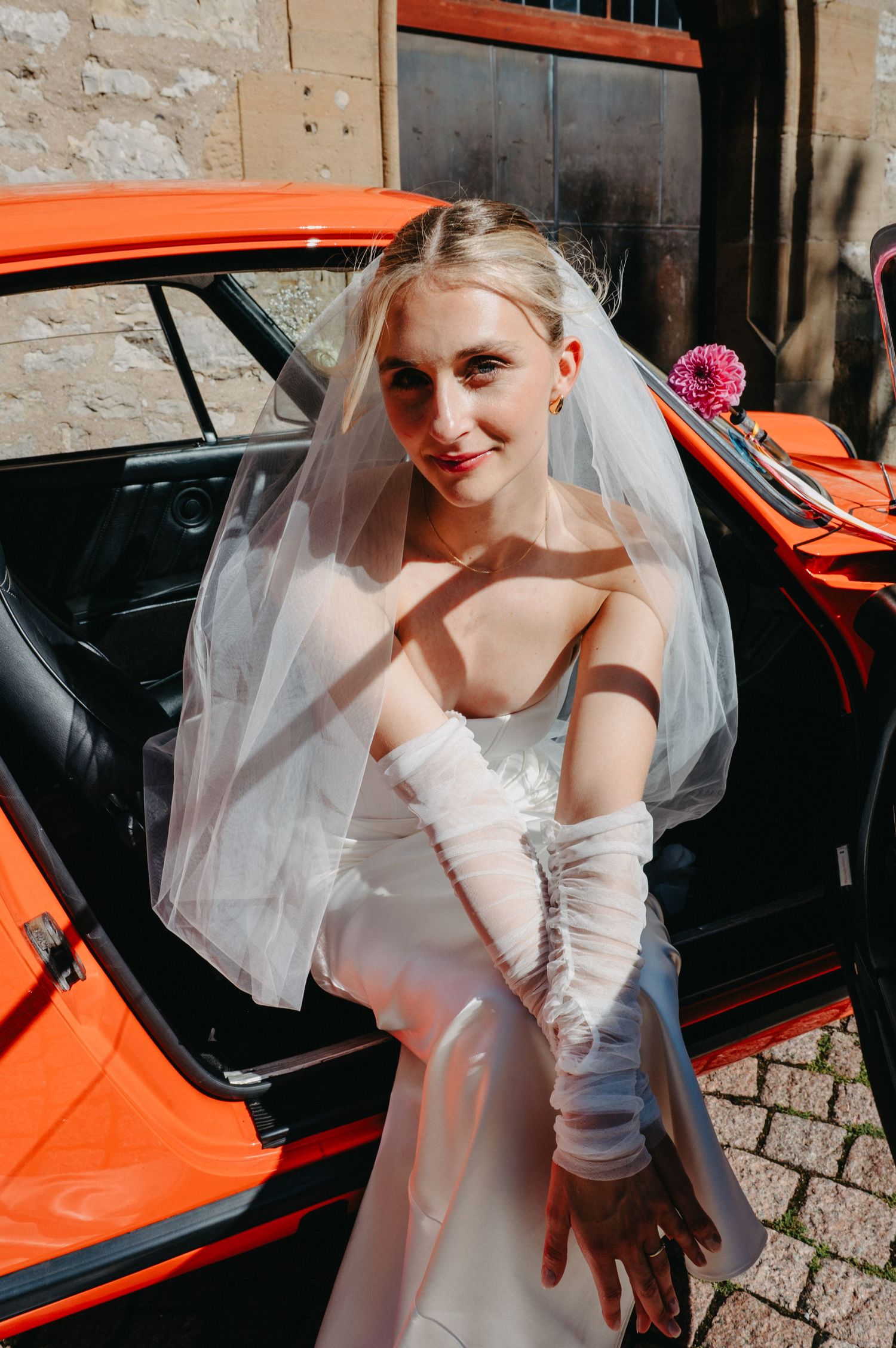 A bride in a white wedding dress and veil poses next to a bright orange vintage car.