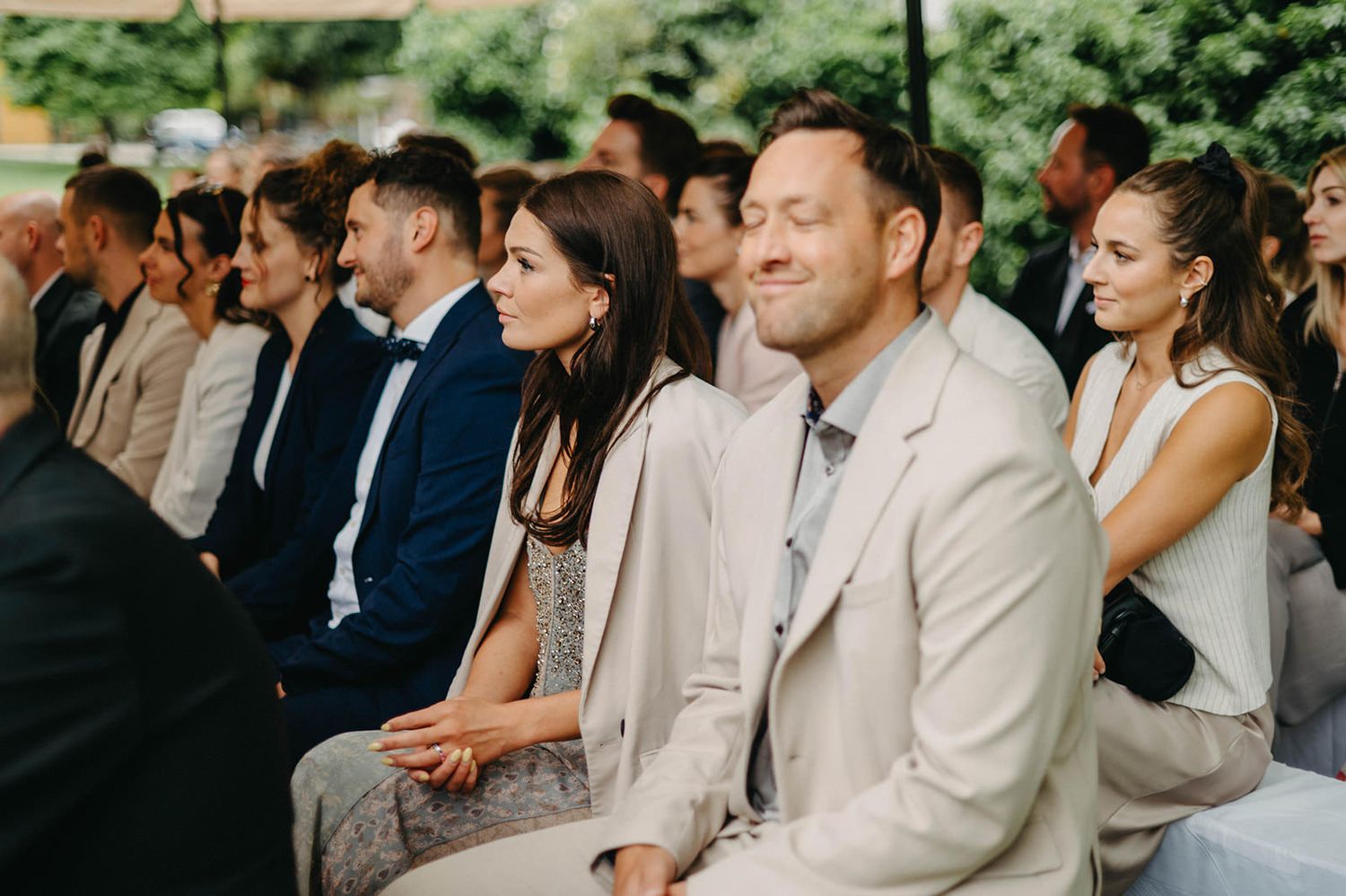 Wedding guests sit together outdoors enjoying a celebration with joyful expressions and laughter.