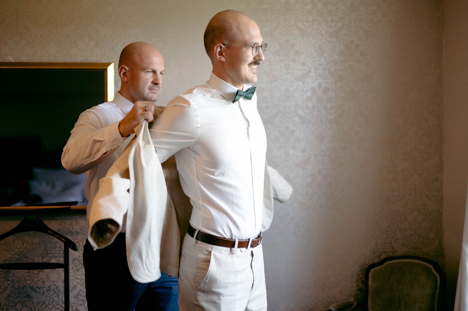 A groom adjusts his formal white dress shirt and bow tie while getting ready for a wedding ceremony.