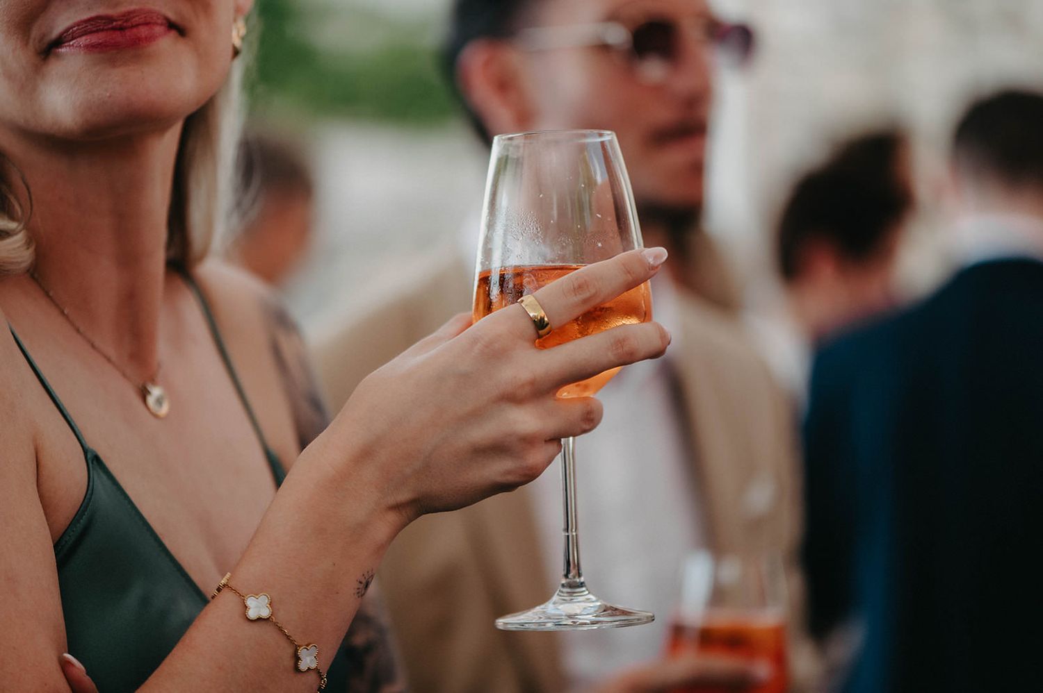 A hand holds a champagne flute filled with rose-colored drink at an elegant outdoor social gathering.