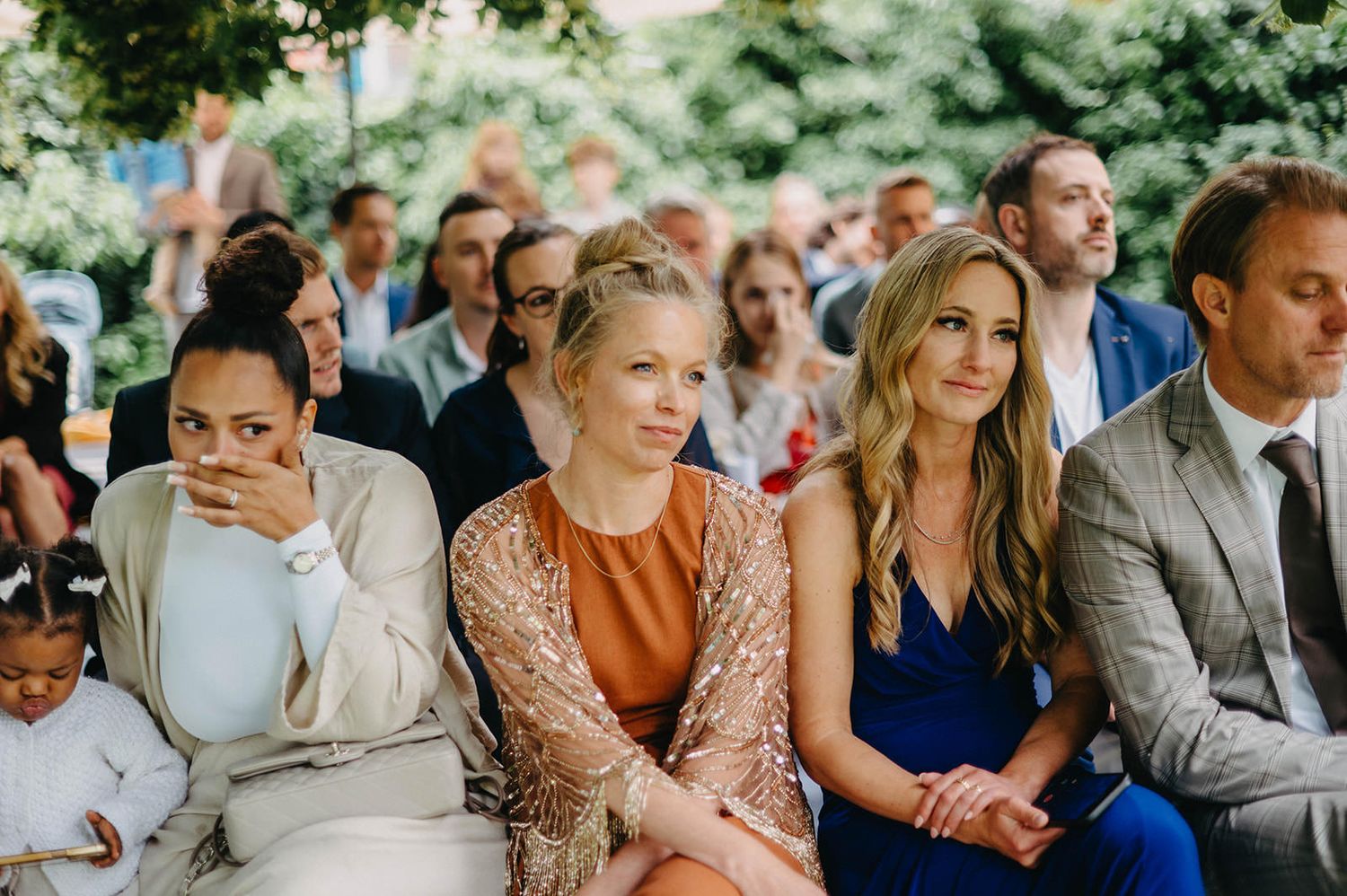 Guests seated outdoors at a wedding ceremony watching with various expressions of emotion and anticipation.