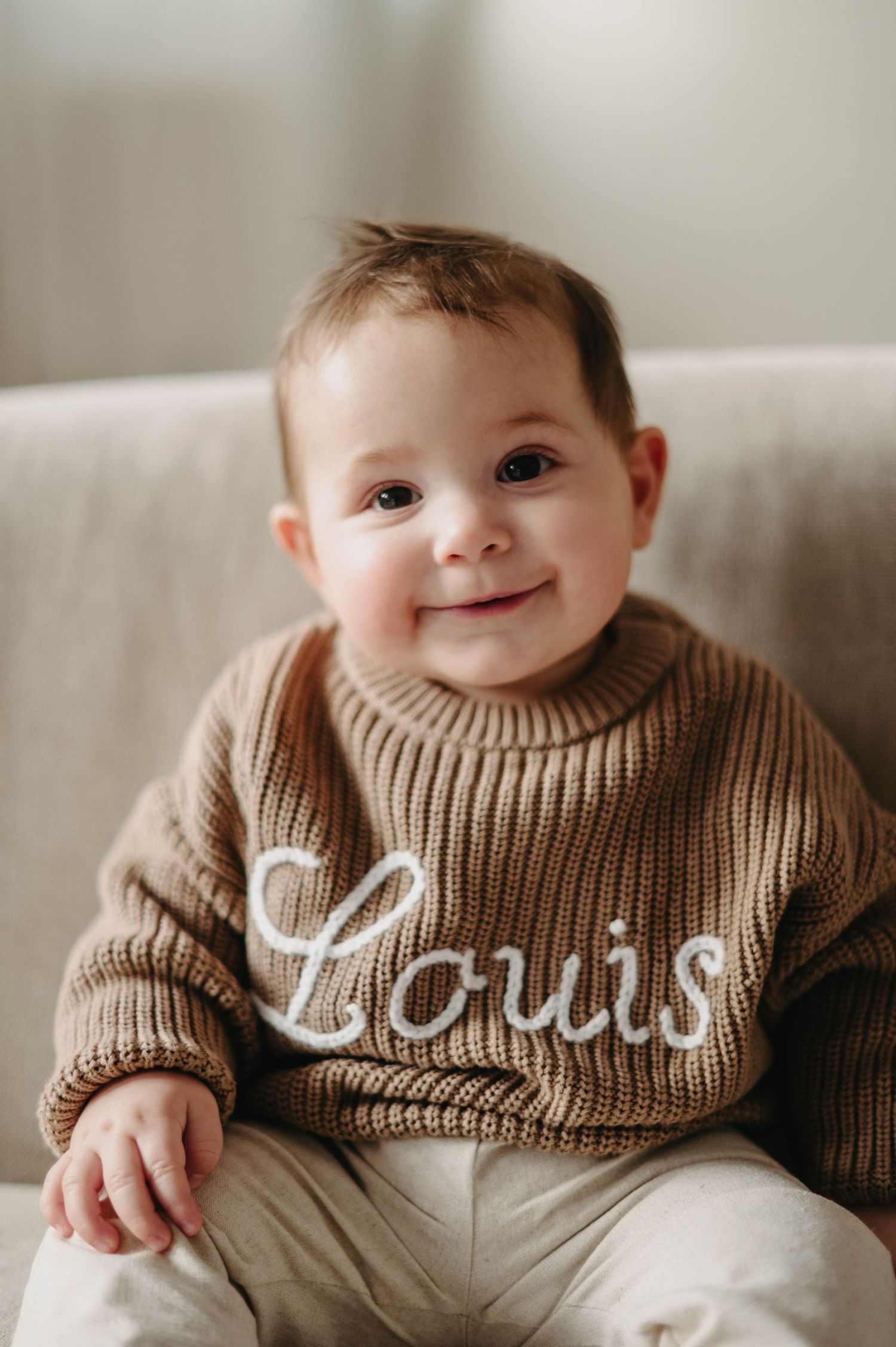 A small child wearing a brown knit sweater with text sits on a light colored couch.