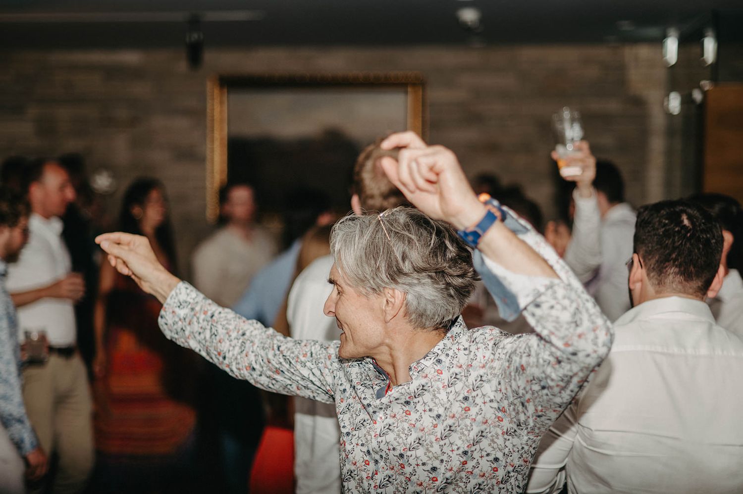 People dancing and celebrating at a wedding reception with a joyful dancer in a floral print dress in the foreground.