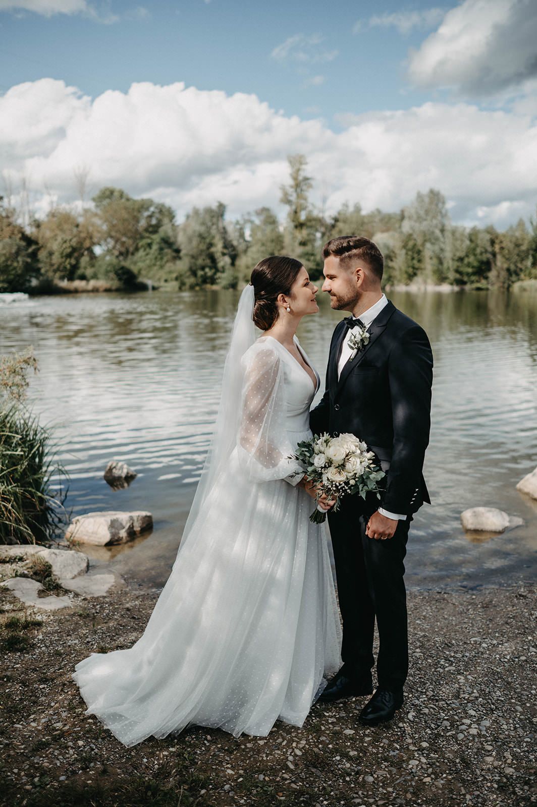 A romantic wedding couple shares intimate moments by a serene lakeside with scattered rocks and trees in the background.