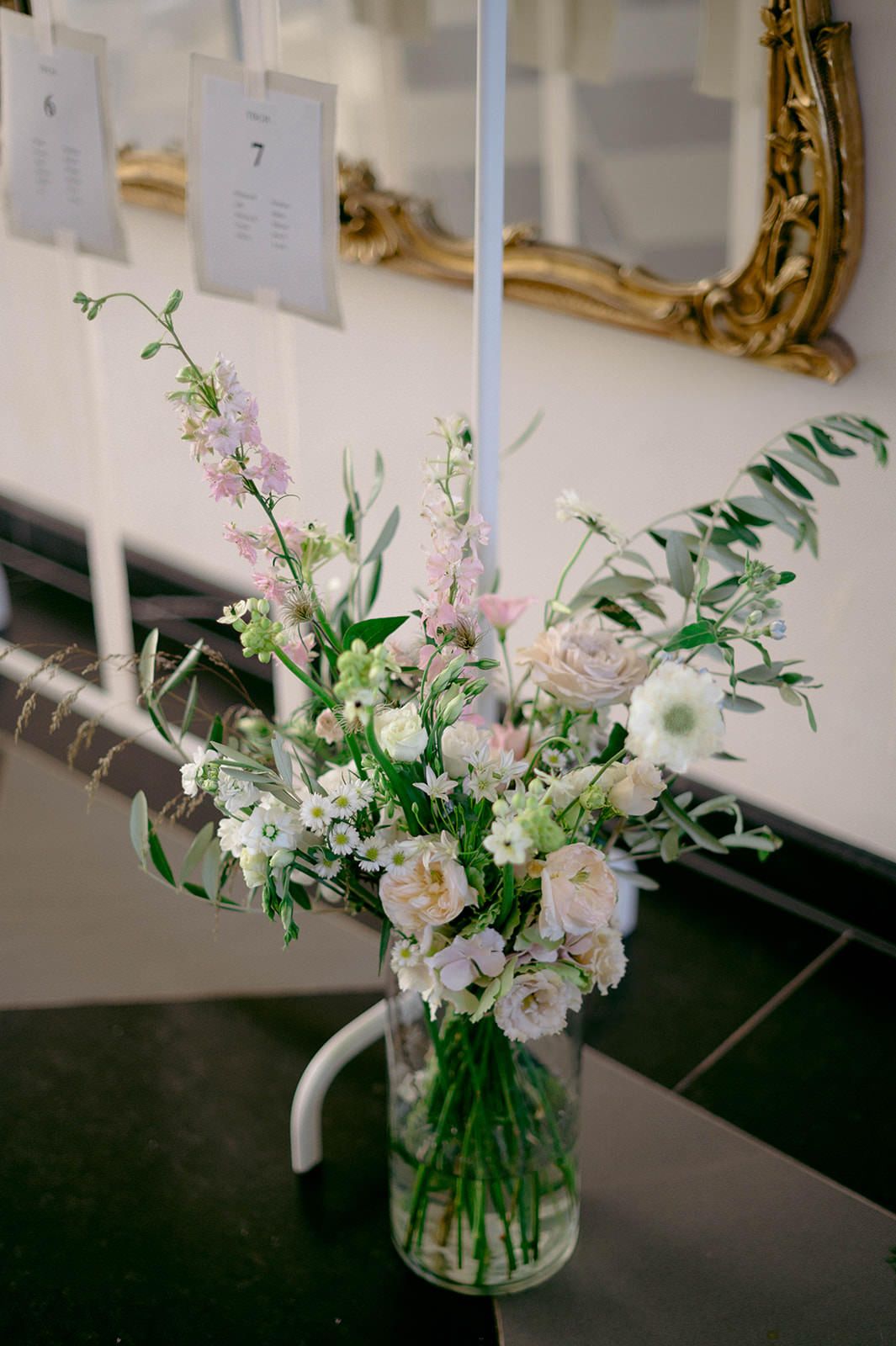 A delicate floral arrangement of white and pink blooms with greenery in a glass vase sits against a white wall.