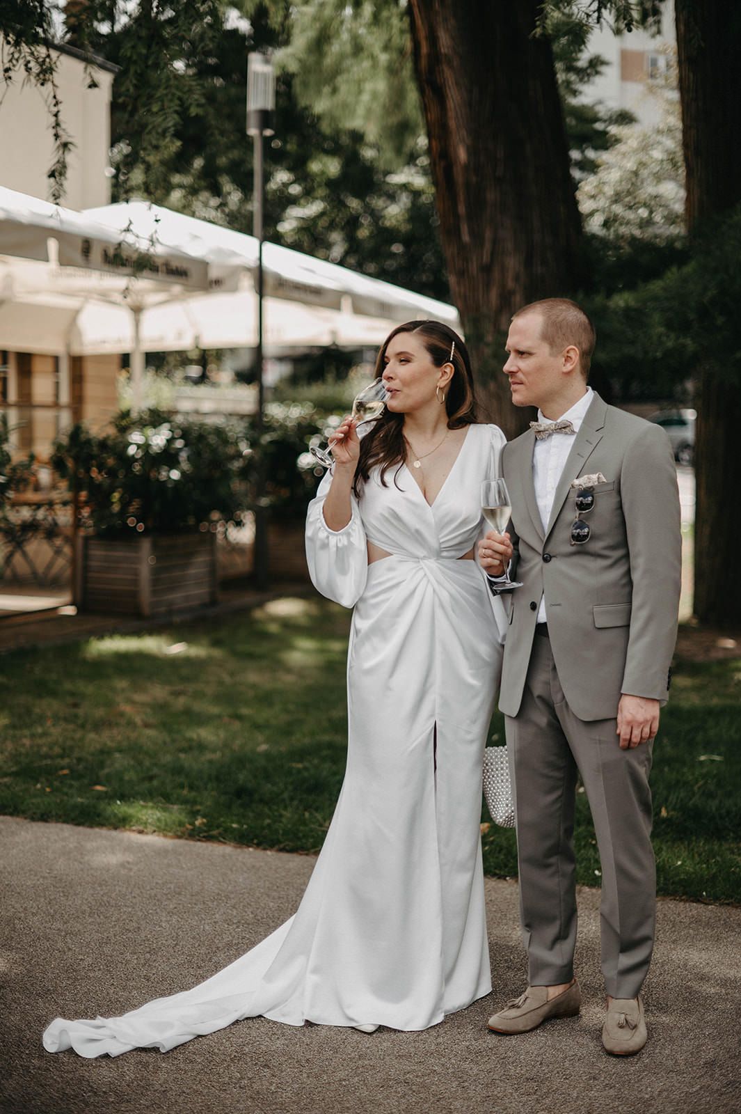 Newlywed couple stands together outdoors, bride in elegant white wrap dress and groom in light gray suit.