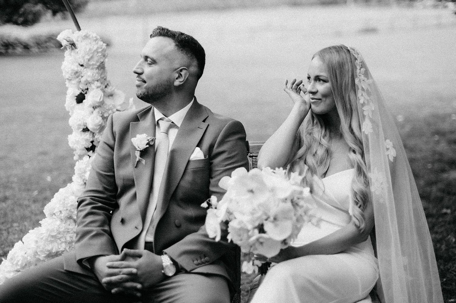 A black and white wedding photograph showing a joyful couple seated outdoors with white floral bouquets.