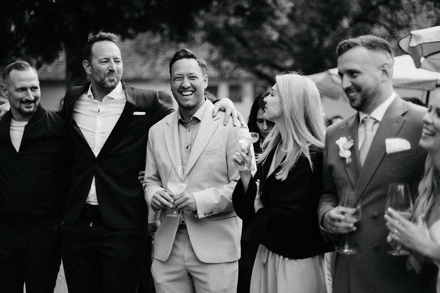 A group of wedding guests laugh and socialize at an outdoor reception celebration.