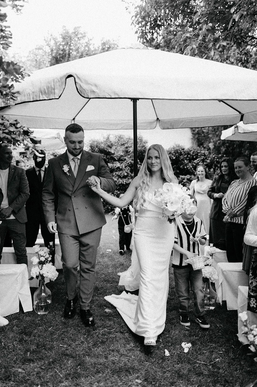 A couple walks under a white umbrella during an outdoor garden wedding ceremony surrounded by guests.