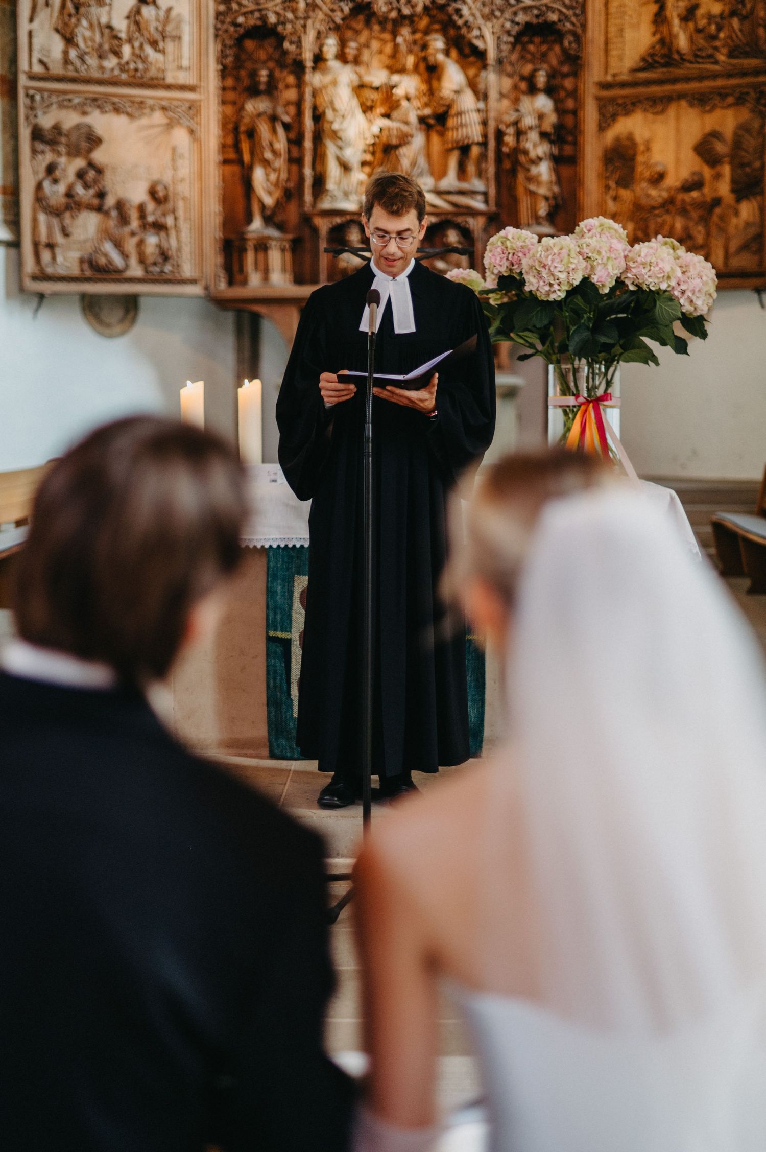 A minister in black robes reads from a book during a wedding ceremony in an ornate church setting.