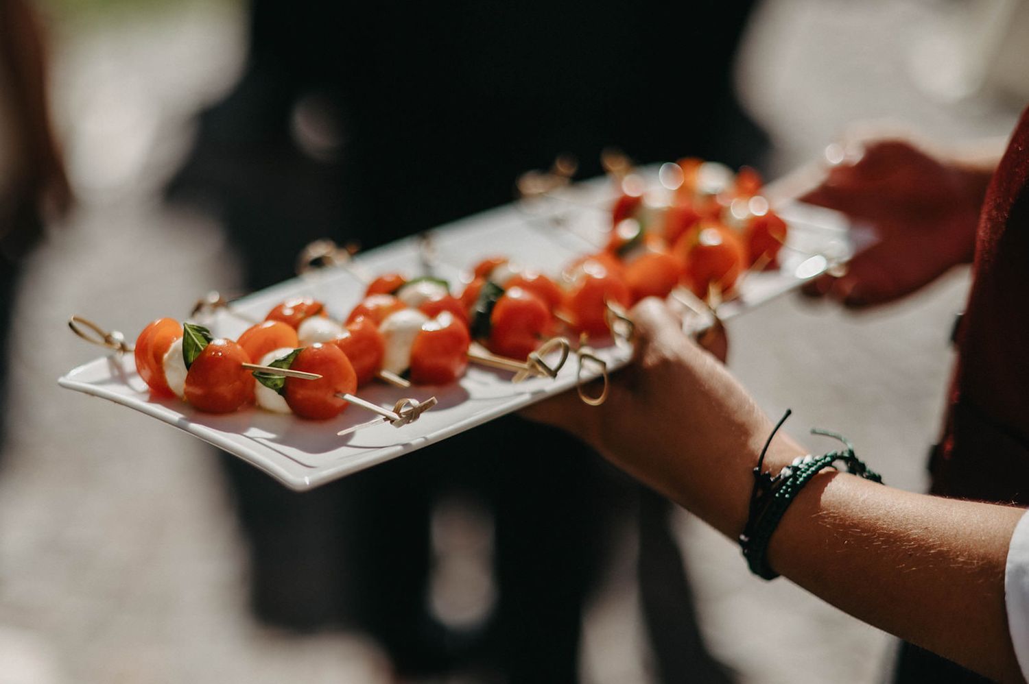 Hands serve skewered cherry tomatoes and mozzarella balls on a white platter at a catering event.