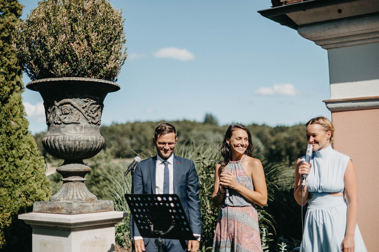 Three people in formal attire stand on a terrace with an ornate stone urn and scenic landscape view in the background.