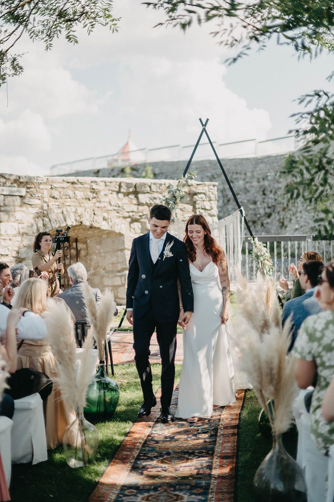 Wedding guests gather outdoors at a rustic stone venue with greenery and string lights creating a romantic atmosphere.