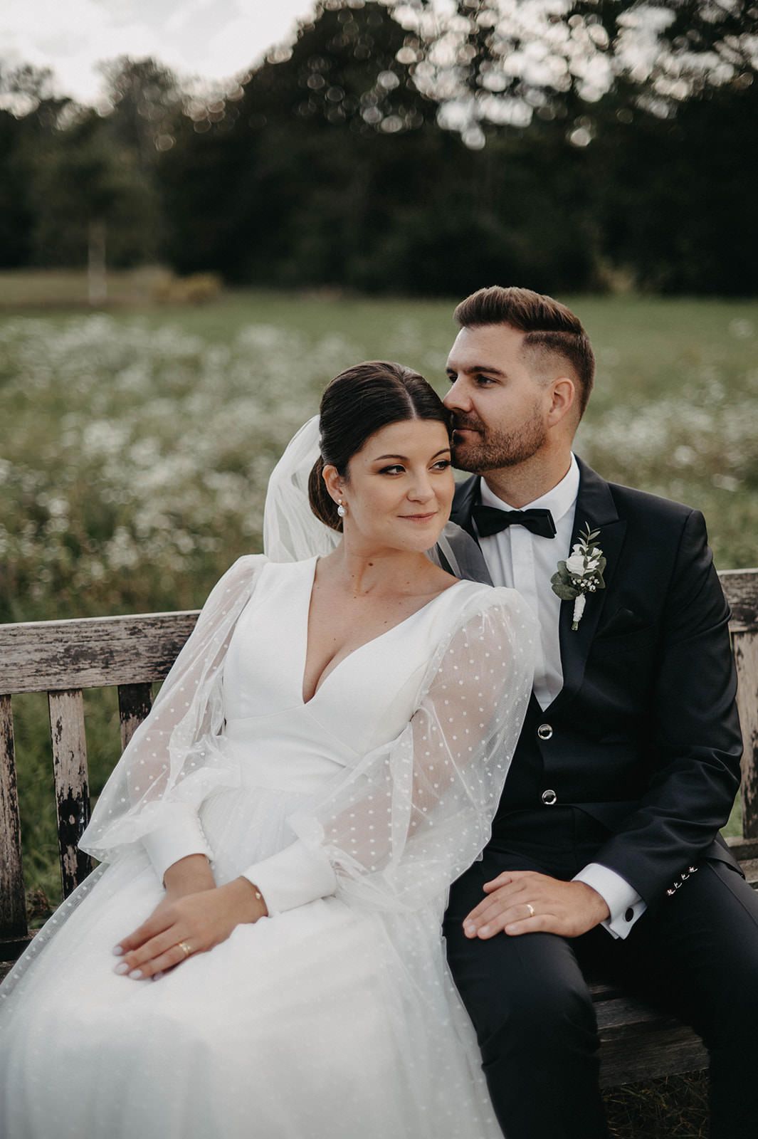 A romantic wedding portrait of a couple sitting on a wooden bench in a wildflower meadow at dusk.