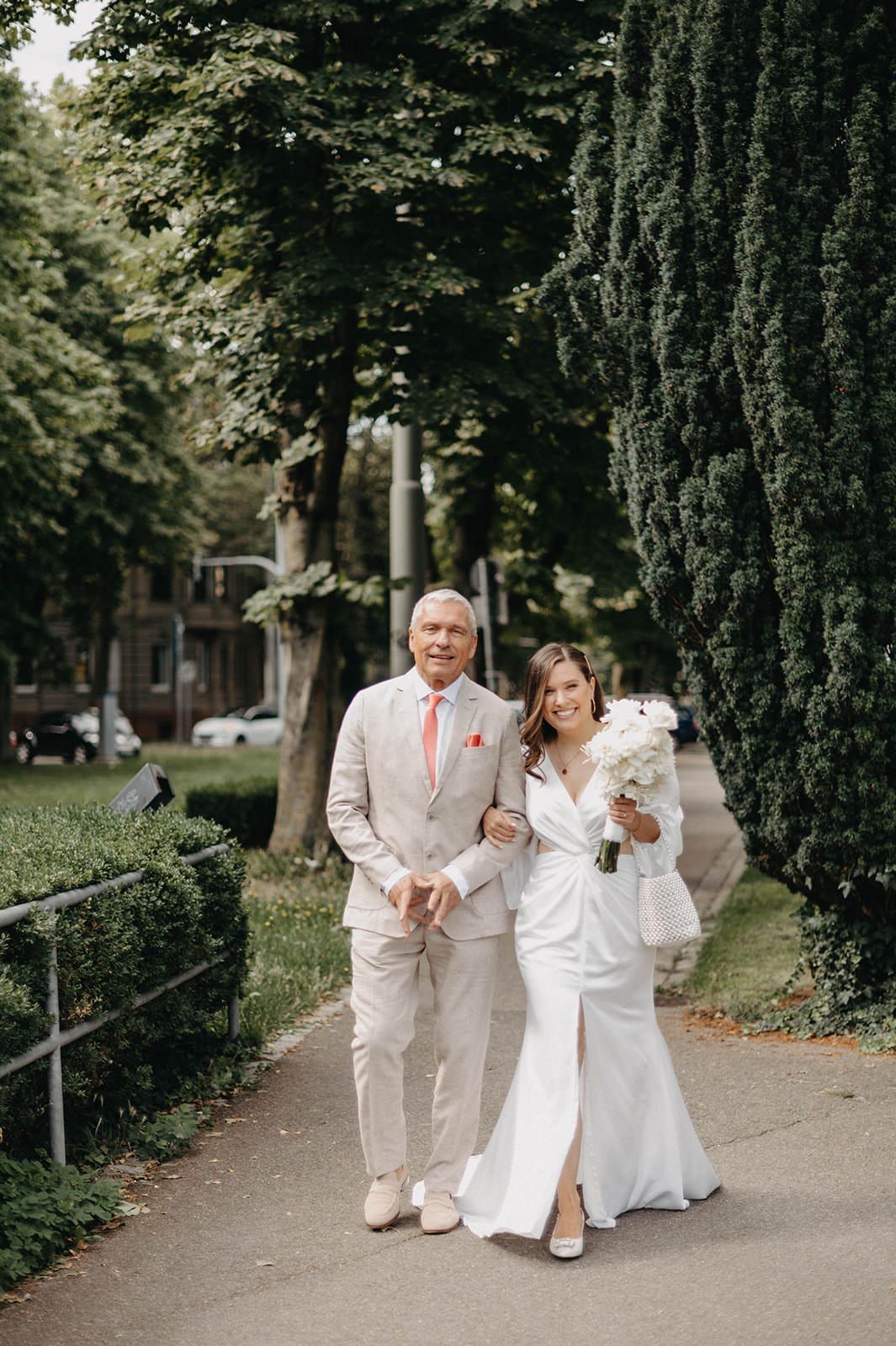A couple walks down a tree-lined path wearing coordinated white and beige wedding attire on their special day.