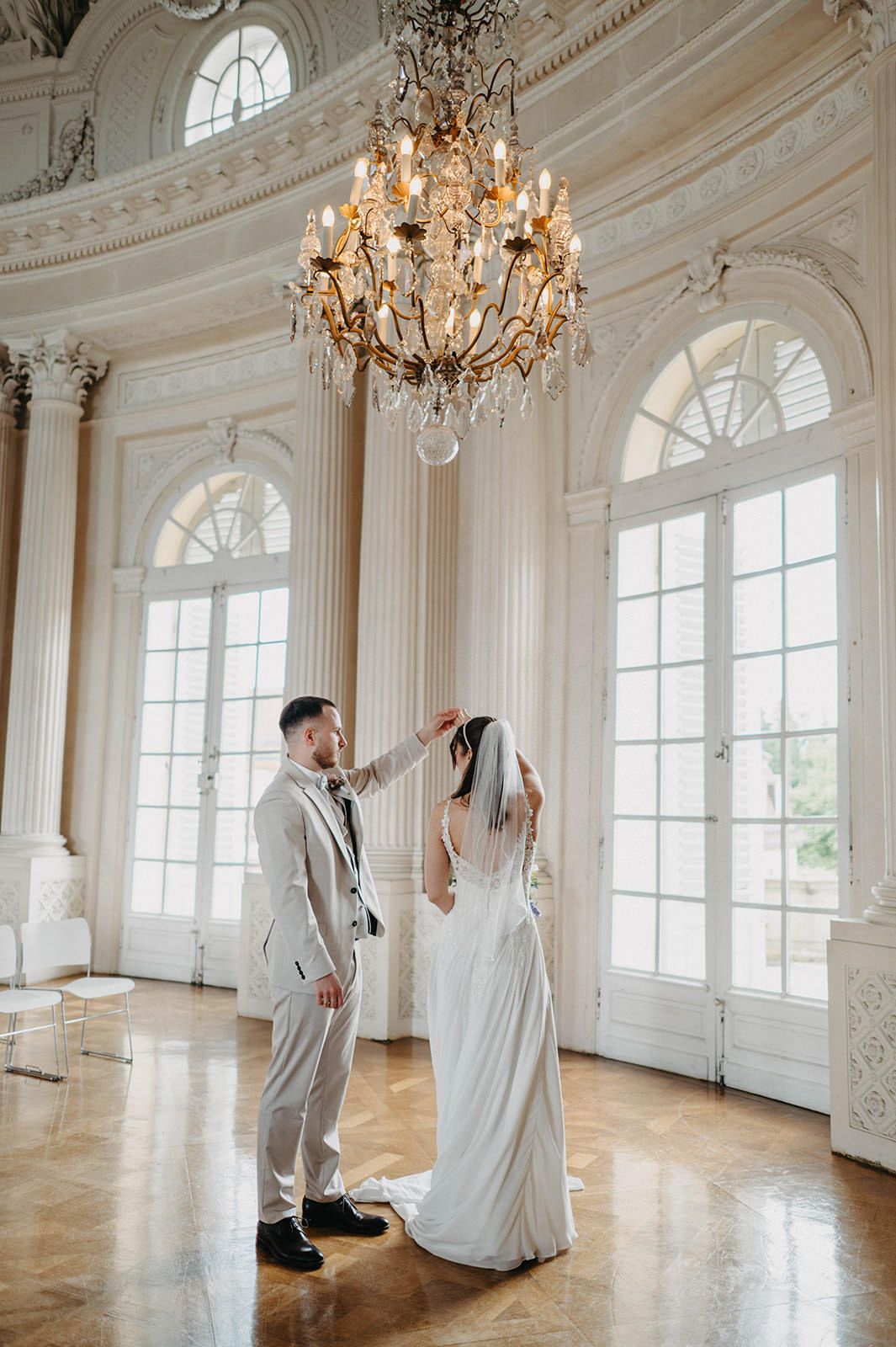 Elegant white ballroom with crystal chandeliers and tall windows showcasing figures in white dresses and gray suits.