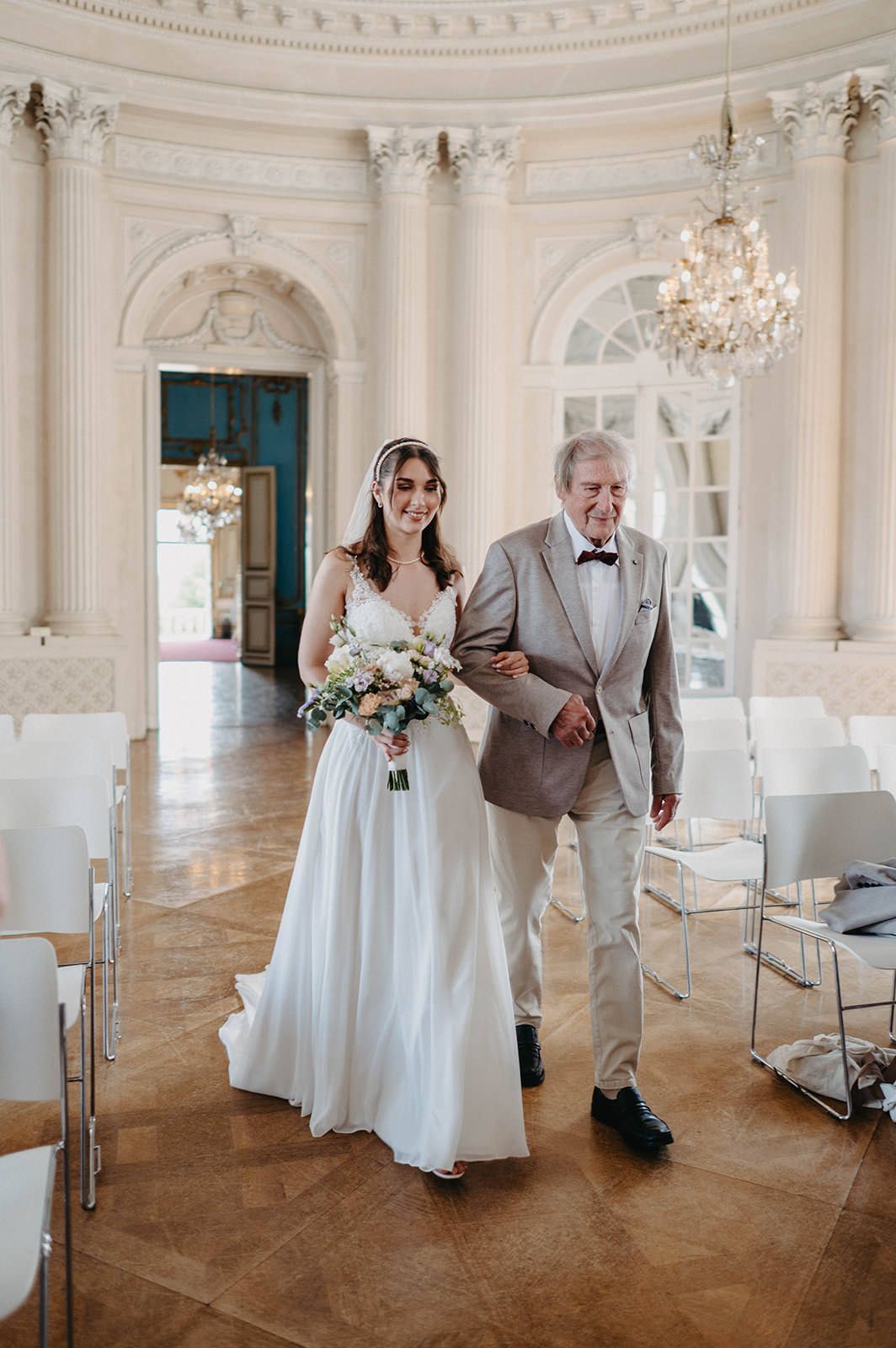 A panoramic view of an elegant white ballroom with chandeliers and a couple celebrating their wedding ceremony.