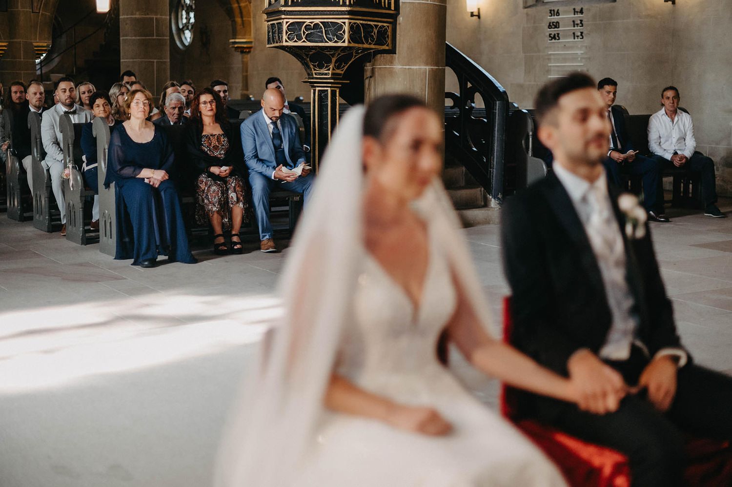 Wedding guests seated in rows at a traditional church ceremony watch as a couple exchanges vows at the altar.