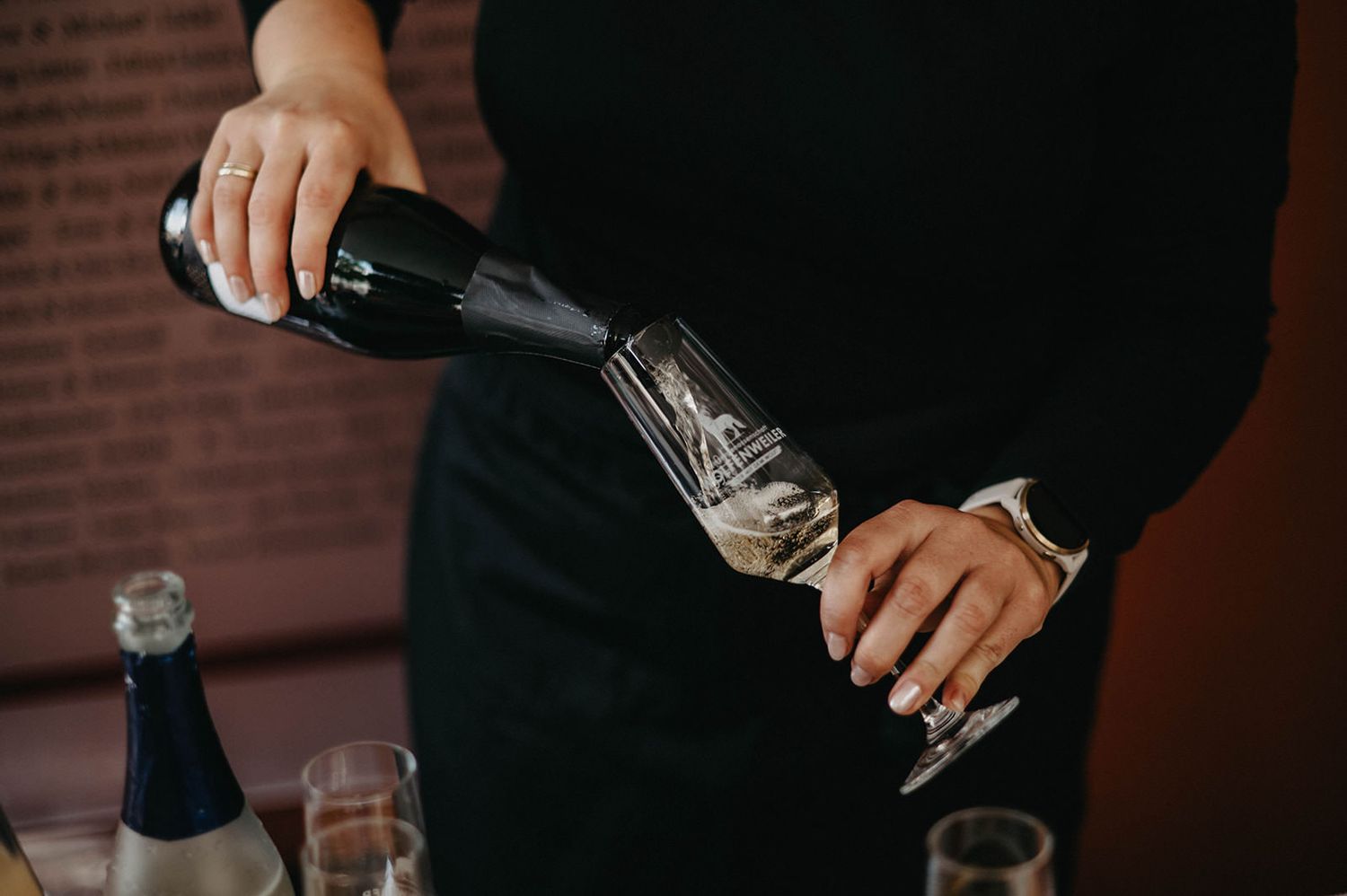 A hand pours a cocktail into a glass at a dimly lit bar with a brick wall backdrop.