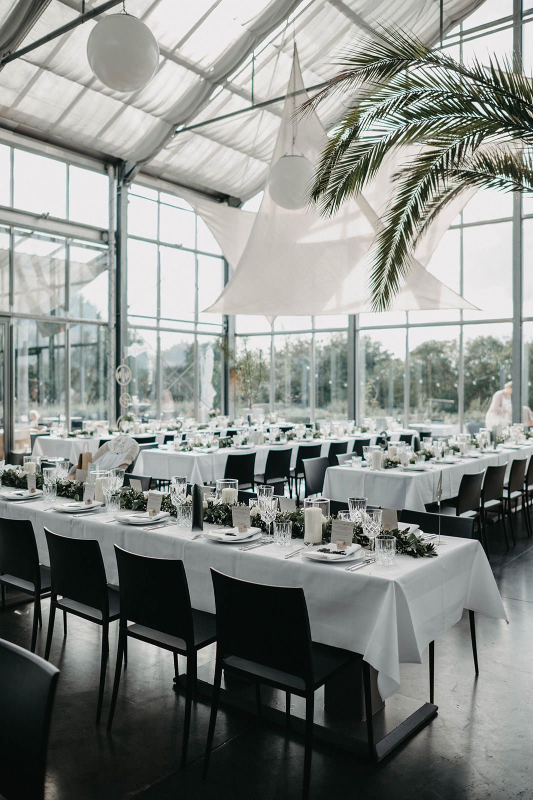 Elegant wedding reception venue with white-clothed tables set up in a modern glass-enclosed space with palm trees.