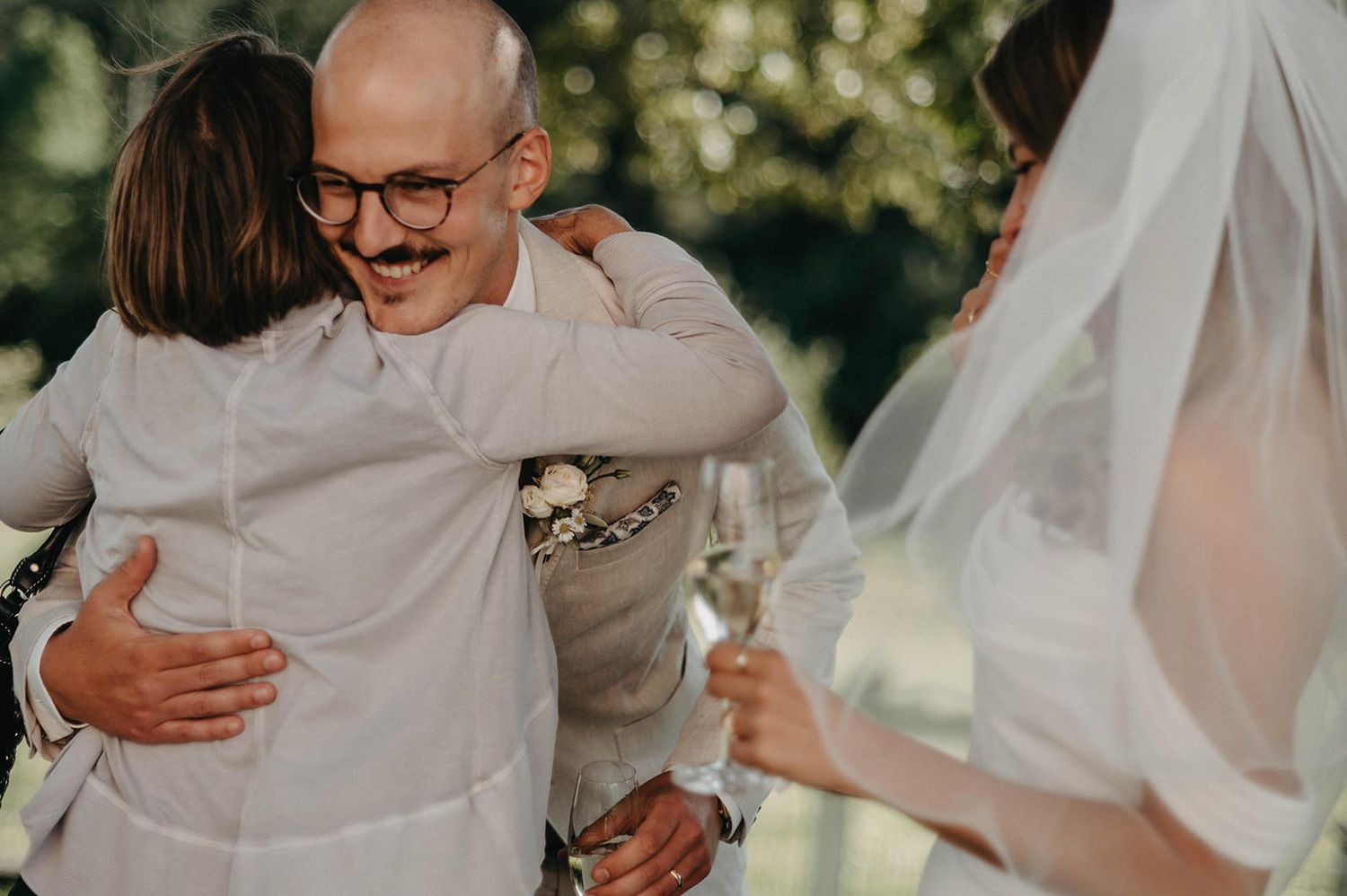 A group of wedding guests share joyful hugs and embraces during an outdoor celebration.