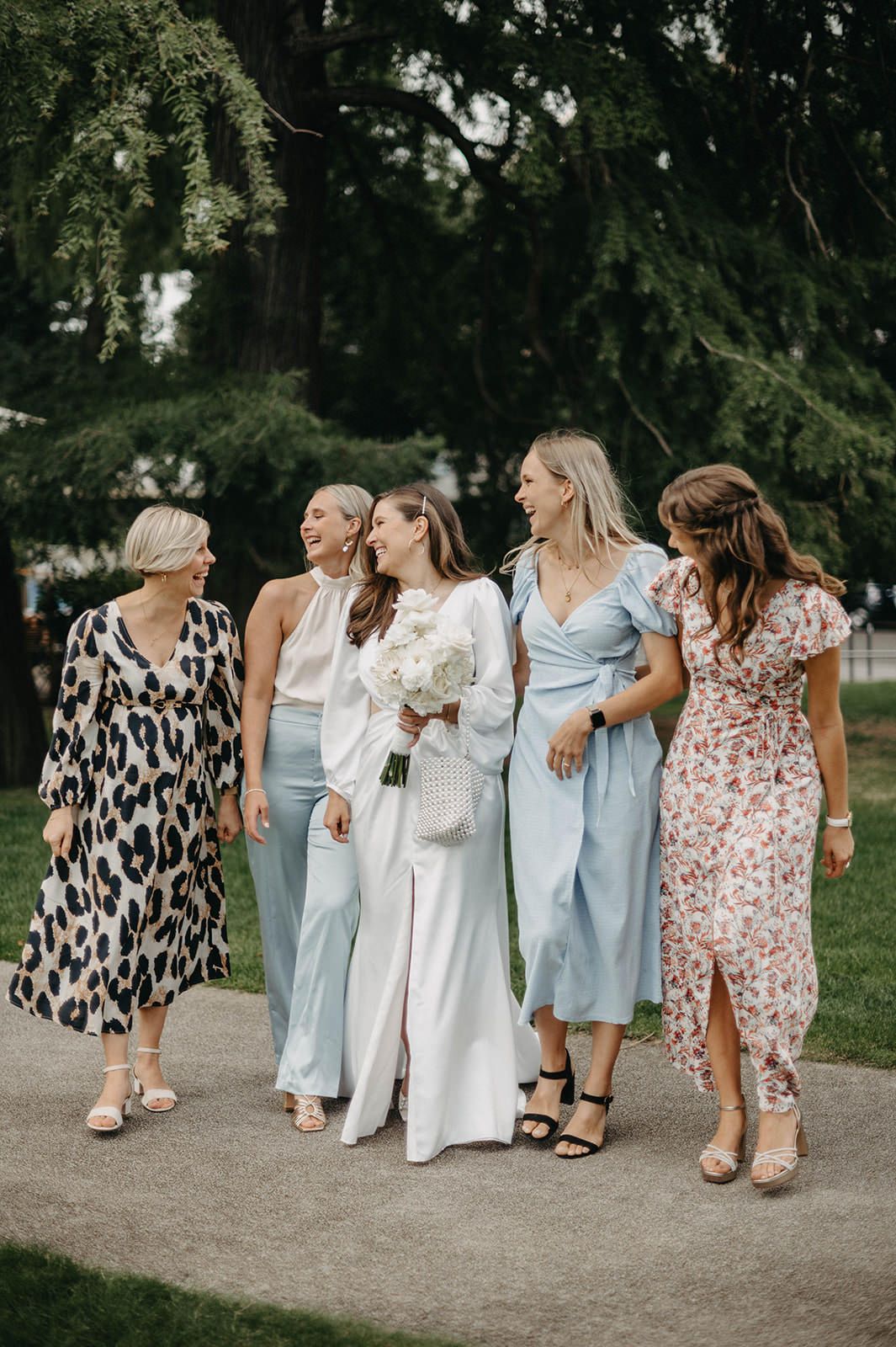 A group of women in light blue dresses and floral prints walk together along a garden path during a summer celebration.