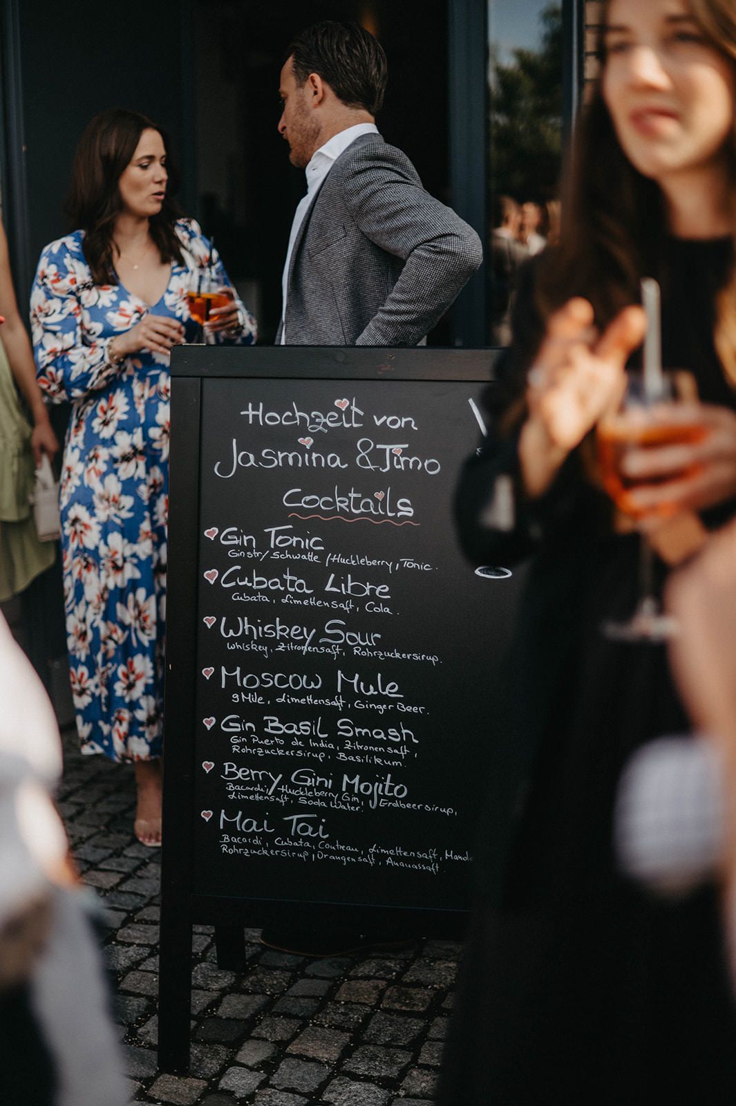 Chalkboard menu displays cocktail offerings at an outdoor event with guests mingling in the background.