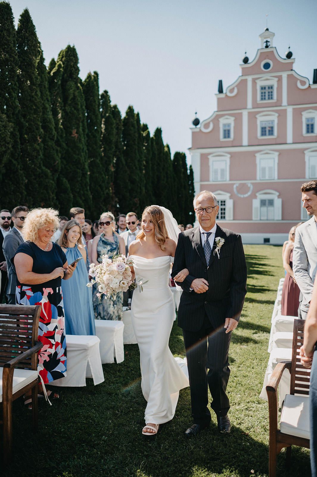 Wedding guests gather outside a pink church building on a sunny day with cypress trees in the background.
