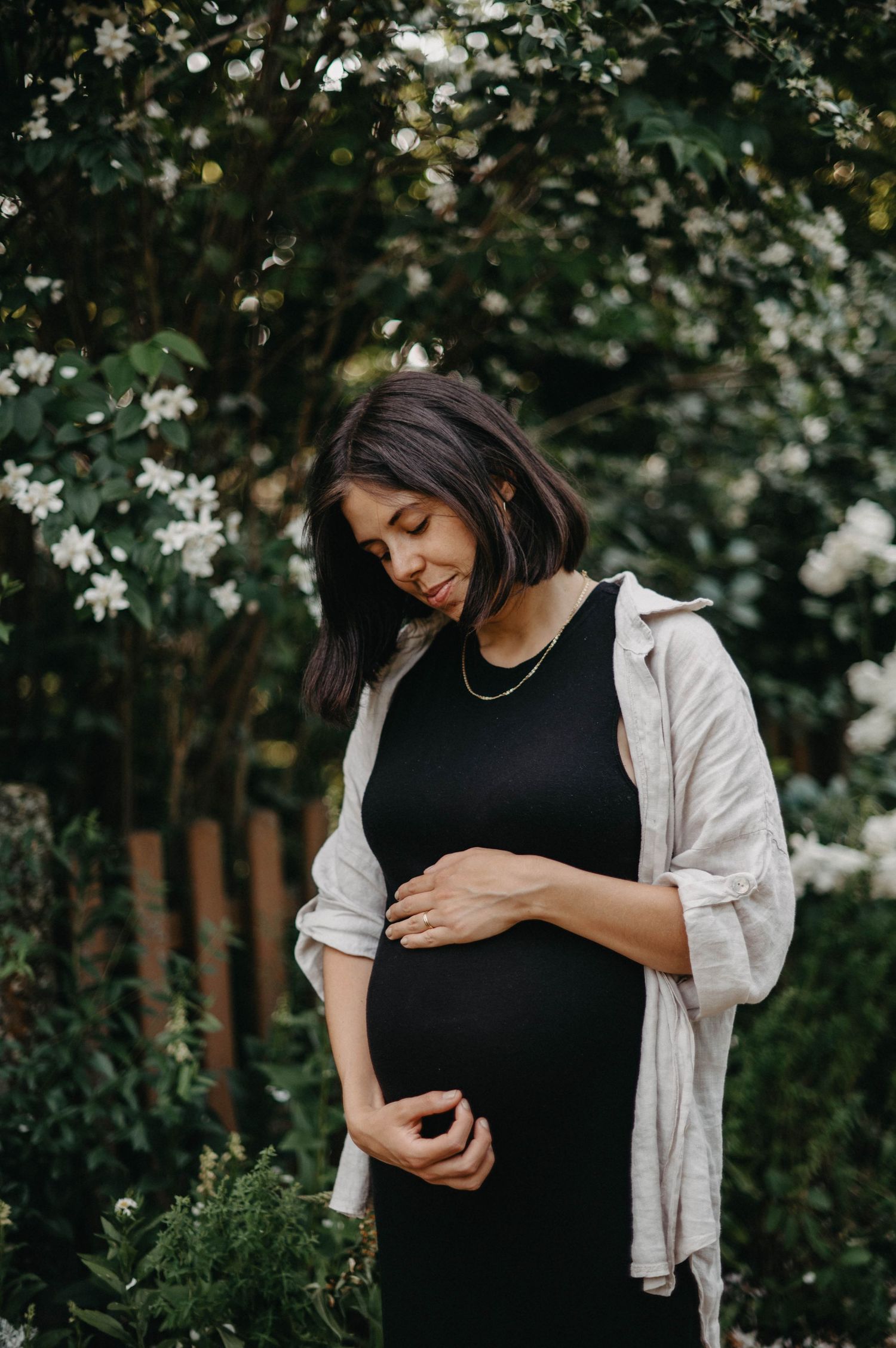 An expectant mother in a black dress touches her belly while standing in a garden with blooming flowers.
