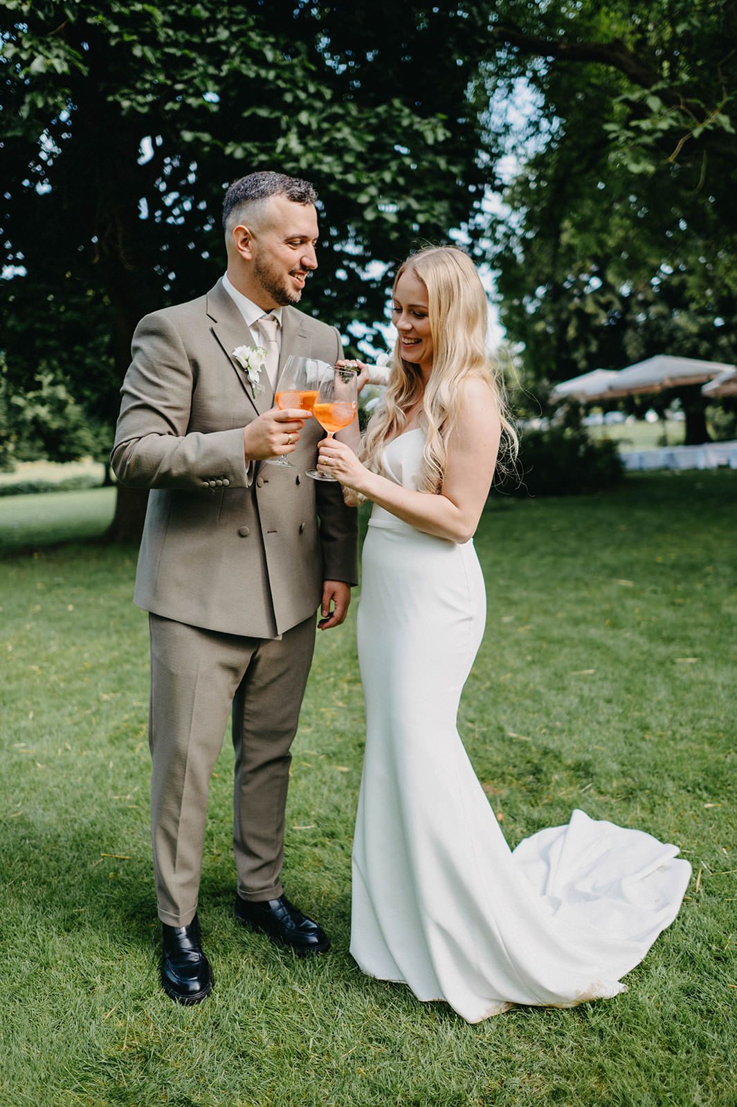 Newlyweds share a toast with orange cocktails on a sunny lawn during their outdoor wedding celebration.