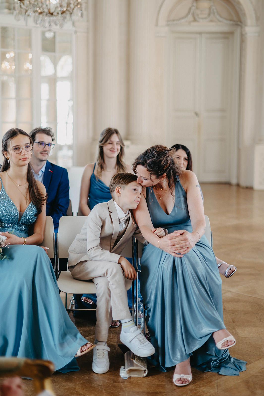 Wedding party members in light blue dresses and suits relax together in an elegant ballroom with white columns.