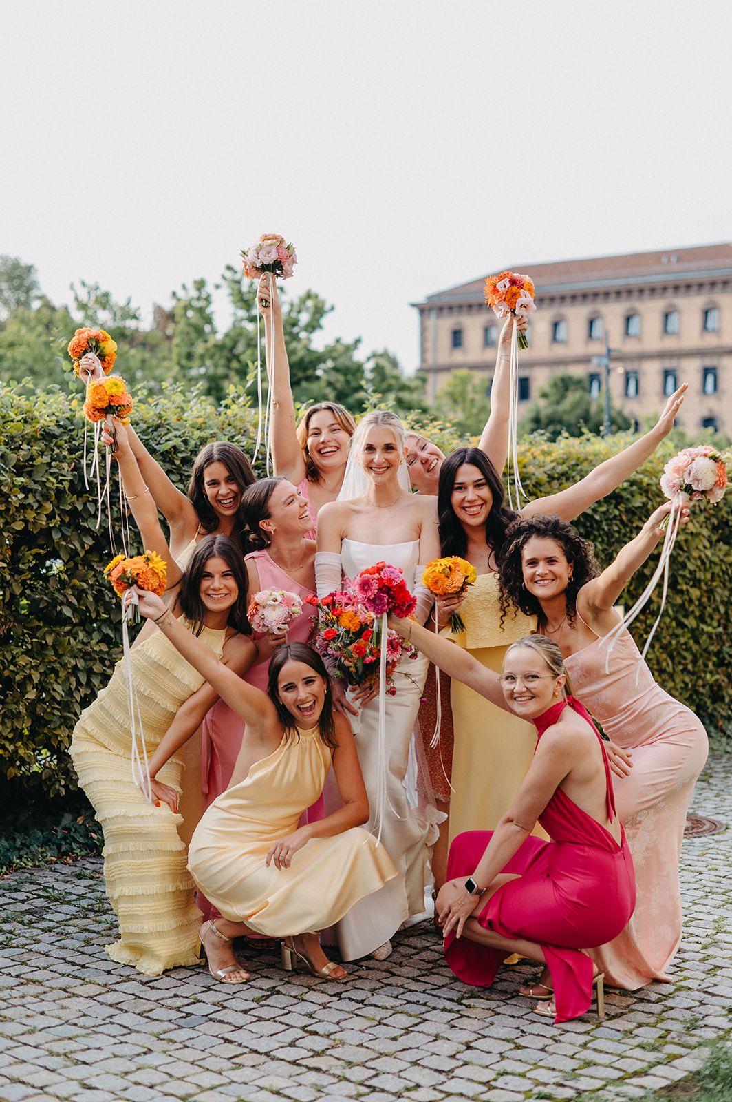 A bridal party in yellow and pink dresses poses joyfully with bouquets on a cobblestone path near a historic building.