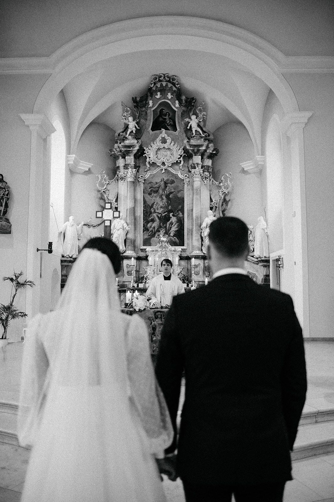A couple stands at the altar of an ornate Catholic church with a baroque-style altarpiece during their wedding ceremony.