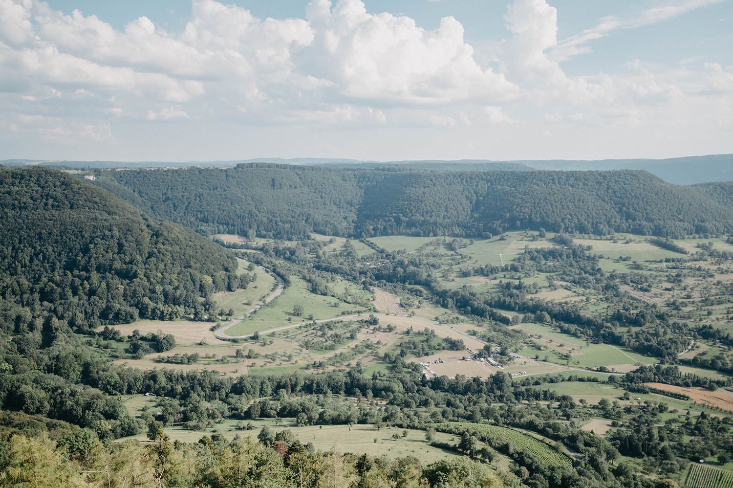 Scenic aerial view of a lush green valley surrounded by forested mountains under a blue summer sky.