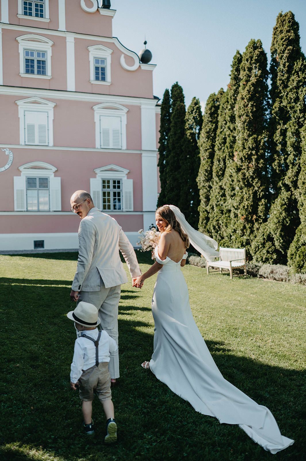 A couple walks together on a sunny lawn in front of a pink mansion with cypress trees in the background.